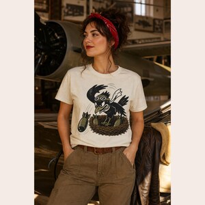 Puede incluir: Una camiseta de manga corta color crema con una ilustraci&oacute;n gr&aacute;fica de un p&aacute;jaro negro con gafas de aviador, posado en un nido y lanzando bombas de color verde oliva. El p&aacute;jaro tiene una expresi&oacute;n decidida y una bufanda blanca que ondea detr&aacute;s.