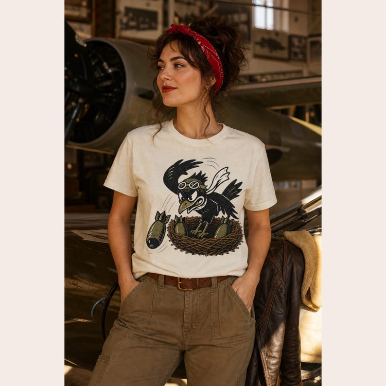 Puede incluir: Una camiseta de manga corta color crema con una ilustraci&oacute;n gr&aacute;fica de un p&aacute;jaro negro con gafas de aviador, posado en un nido y lanzando bombas de color verde oliva. El p&aacute;jaro tiene una expresi&oacute;n decidida y una bufanda blanca que ondea detr&aacute;s.