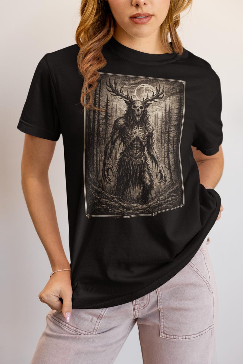 May include: A black short-sleeved t-shirt featuring a rectangular graphic print of a skeletal creature with large antlers standing in a dark forest under a full moon. The illustration is rendered in shades of brown, black, and white.