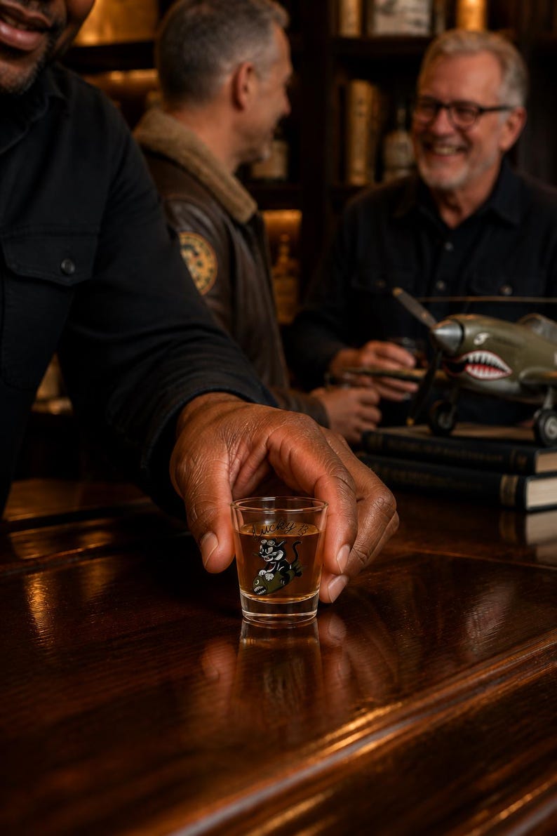 May include: A clear glass shot glass filled with an amber liquid, featuring a graphic of a black cat riding a green military-style bomb with the word Lucky written in cursive script above the illustration, positioned on a dark wooden surface.