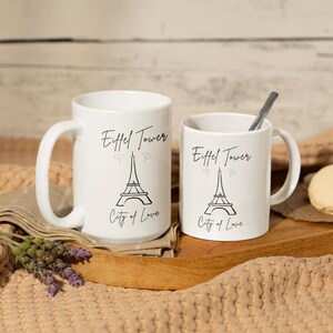 May include: Two white ceramic mugs with black text and illustrations. The mugs feature the Eiffel Tower and the words "Eiffel Tower" and "City of Love". One mug is larger than the other. A spoon rests in the smaller mug.