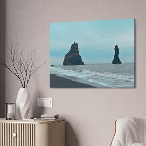 May include: A canvas print depicting a coastal landscape with two large, dark rock formations rising from the ocean. The scene features a black sand beach, foamy waves, and a muted blue sky, creating a serene and natural aesthetic.
