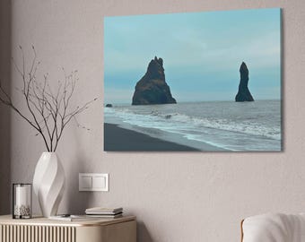 May include: A canvas print depicting a coastal landscape with two large, dark rock formations rising from the ocean. The scene features a black sand beach, foamy waves, and a muted blue sky, creating a serene and natural aesthetic.