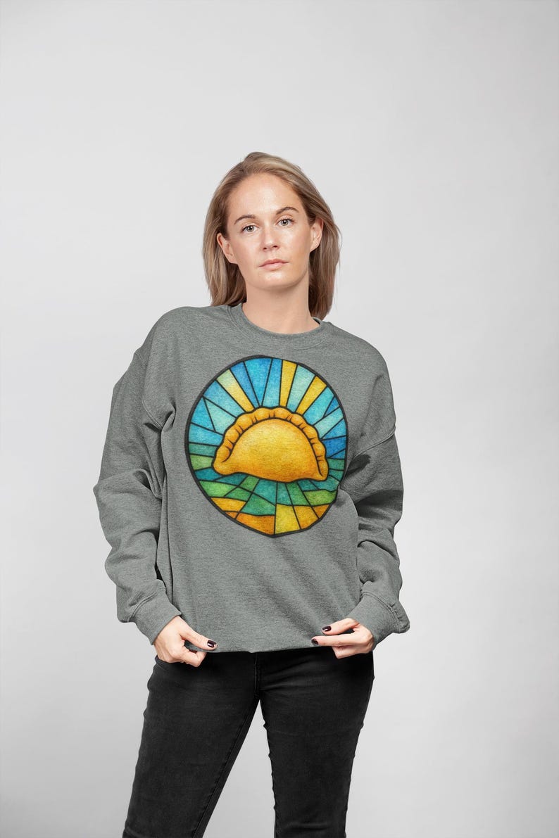 May include: A gray sweatshirt featuring a colorful graphic of a golden empanada in a stained-glass style circle. The design includes blue, yellow, and green hues. The sweatshirt is worn by a person in black jeans.