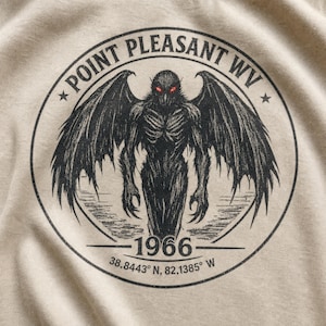 May include: A beige t-shirt featuring a black circular graphic of a winged creature with glowing red eyes. The text reads POINT PLEASANT WV, 1966, and 38.8443 N, 82.1385 W. The design is centered on the fabric with a detailed, sketch-like illustration.