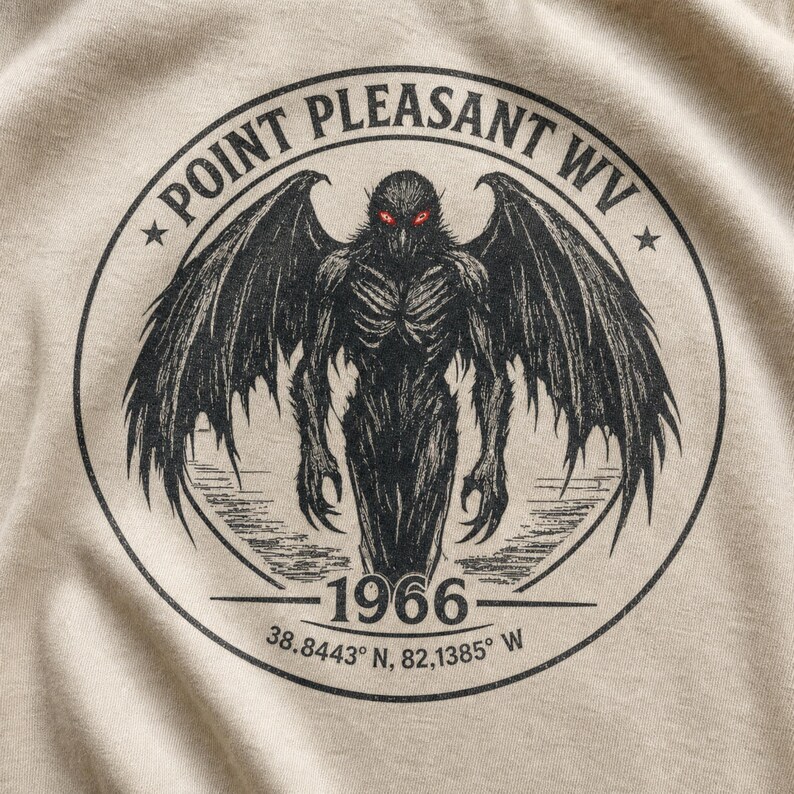 May include: A beige t-shirt featuring a black circular graphic of a winged creature with glowing red eyes. The text reads POINT PLEASANT WV, 1966, and 38.8443 N, 82.1385 W. The design is centered on the fabric with a detailed, sketch-like illustration.