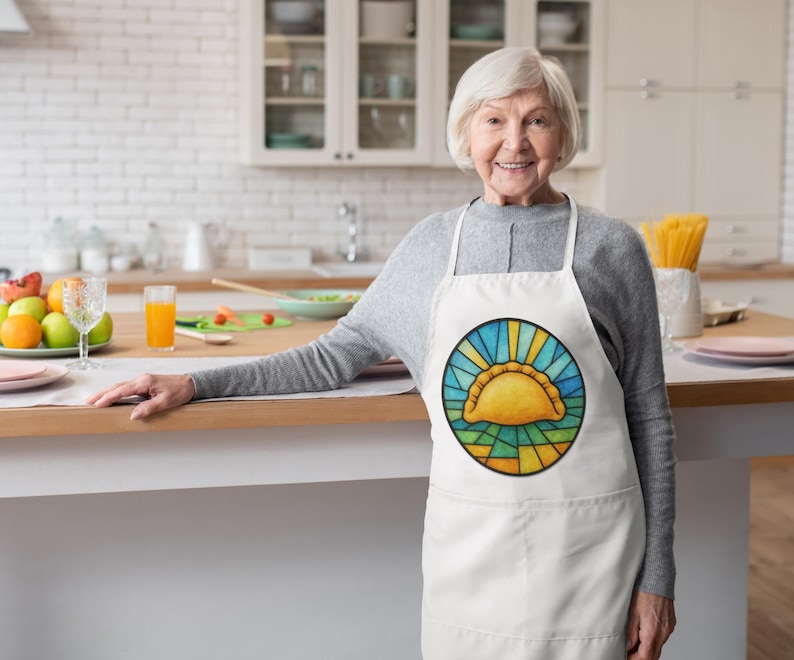 May include: White apron with a colorful, circular design featuring a yellow empanada-like pastry. The design is framed by blue and yellow rays, resembling stained glass. The apron has a pocket and is worn by a person in a kitchen setting.