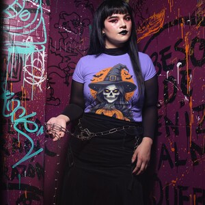 May include: A person wearing a purple t-shirt with a Halloween witch skull graphic, black skirt, and chain belt. The background is a graffiti-covered wall with purple and green paint.
