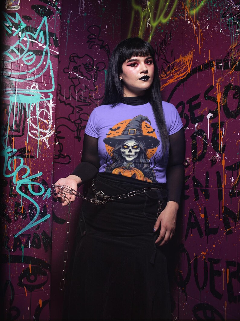 May include: A person wearing a purple t-shirt with a Halloween witch skull graphic, black skirt, and chain belt. The background is a graffiti-covered wall with purple and green paint.