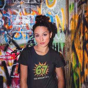 May include: Black t-shirt with the number "67" in green and yellow, and the text "YOU WOULDN'T GET IT." The background is a graffiti-covered wall with various colors and designs.