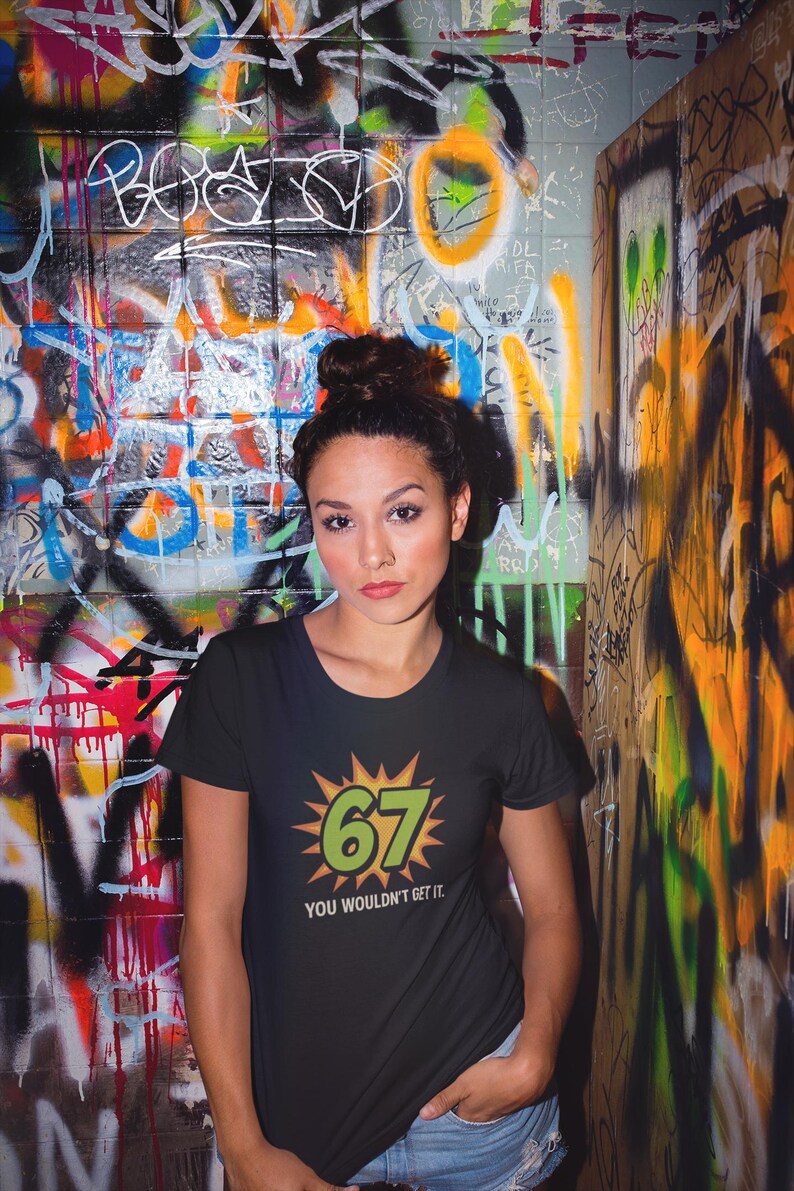May include: Black t-shirt with the number "67" in green and yellow, and the text "YOU WOULDN'T GET IT." The background is a graffiti-covered wall with various colors and designs.