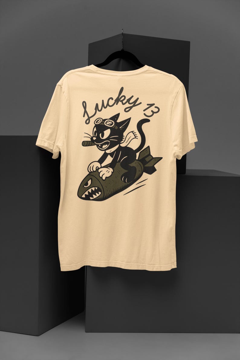 May include: Cream-colored t-shirt featuring a cartoon cat riding a bomb, with the text "Lucky 13." The black cat wears goggles and smokes a cigar. The bomb has a menacing face. The shirt is displayed on a black hanger.