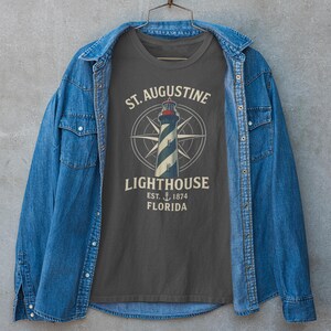 May include: A dark gray t-shirt featuring a lighthouse design with the text "St. Augustine Lighthouse, Est. 1874, Florida." The shirt is displayed on a hanger, framed by a blue denim jacket.