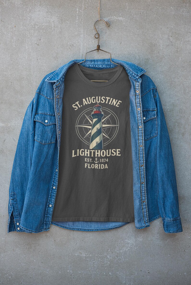 May include: A dark gray t-shirt featuring a lighthouse design with the text "St. Augustine Lighthouse, Est. 1874, Florida." The shirt is displayed on a hanger, framed by a blue denim jacket.