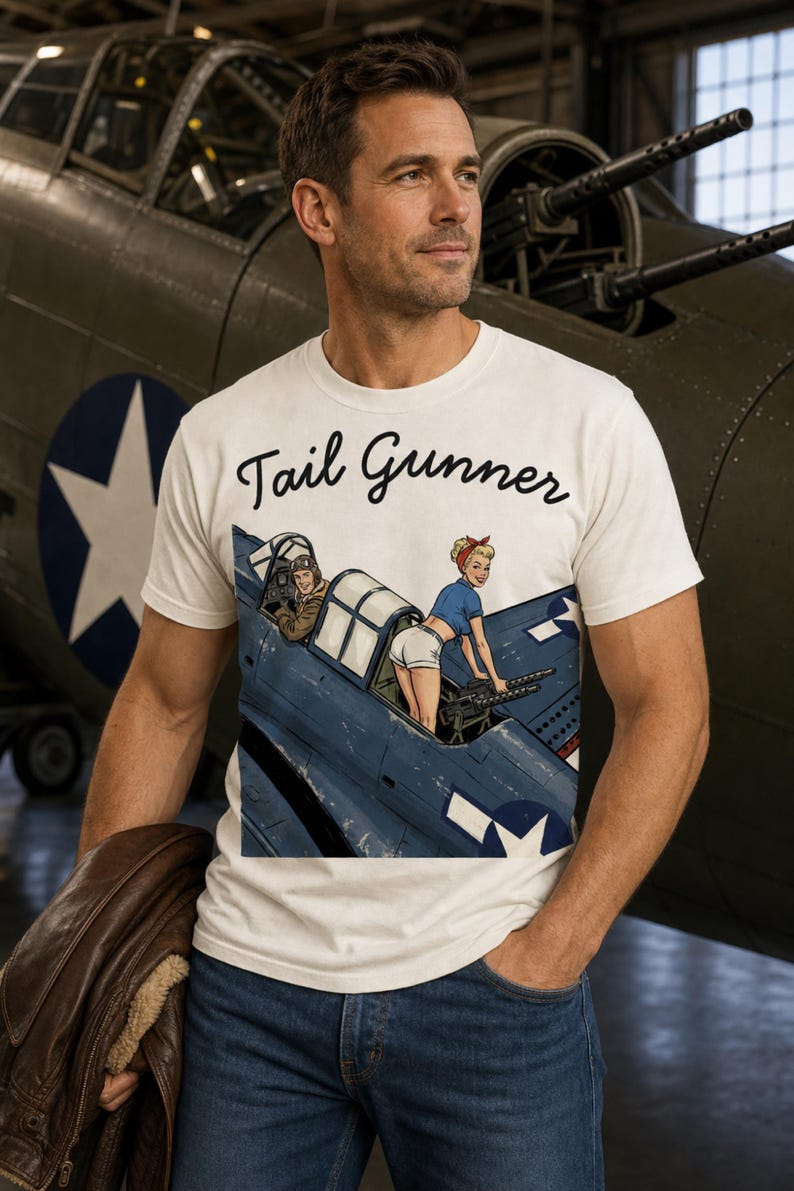 May include: A cream-colored short-sleeve t-shirt featuring the text Tail Gunner in black script above a graphic illustration of a blue military aircraft with a pilot and a person operating a machine gun from the rear cockpit area.