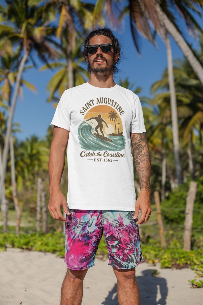 May include: White t-shirt with a graphic of a surfer on a wave, the text "SAINT AUGUSTINE" and "Catch the Coastline - EST. 1565". The person is wearing colorful shorts. Palm trees are in the background.