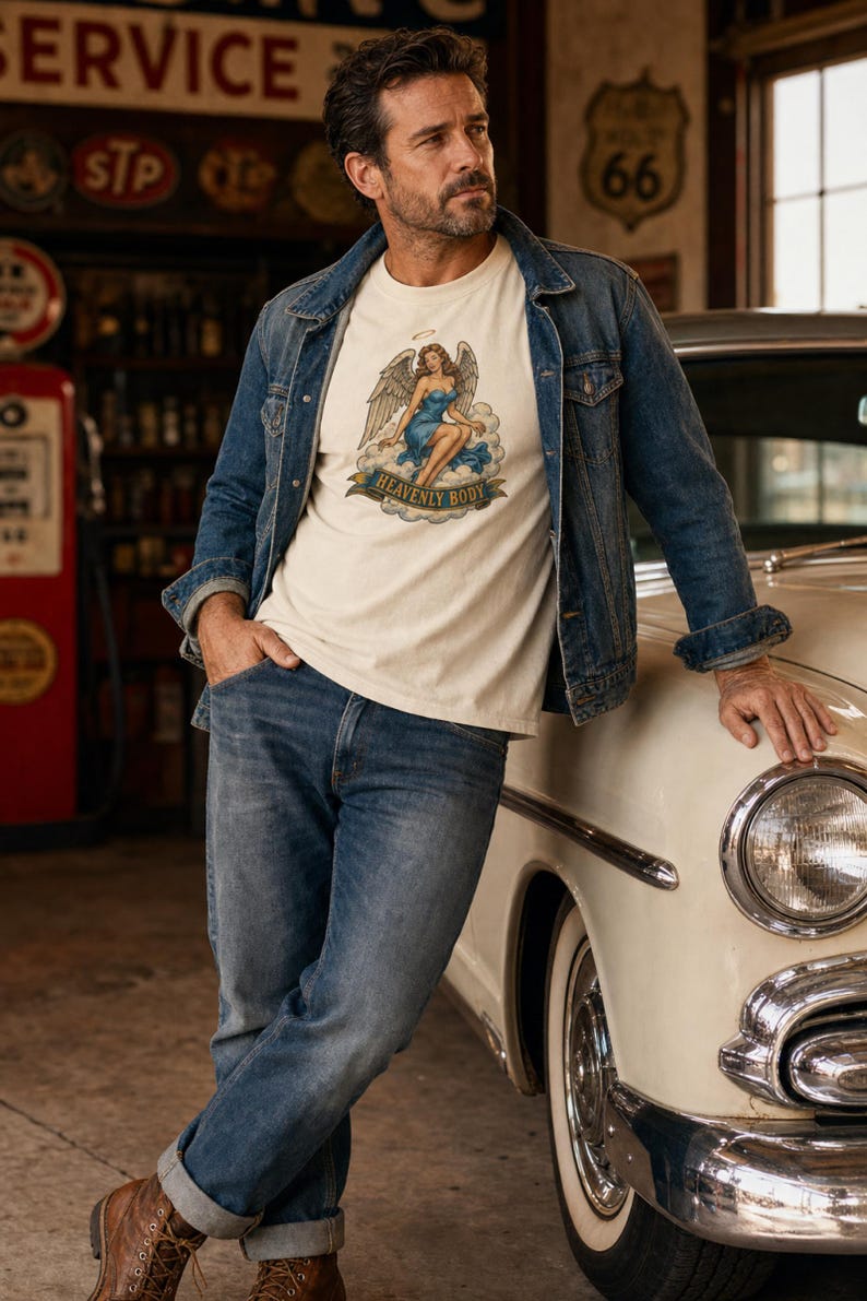 May include: A cream-colored short-sleeve t-shirt featuring a graphic print of a winged figure sitting on a cloud with a halo above their head. Below the figure, a banner displays the text HEAVENLY BODY in dark blue capital letters.