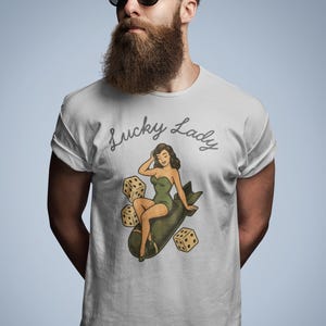 May include: A light gray short-sleeved t-shirt featuring a graphic print of a woman in a green swimsuit sitting on a dark green bomb, surrounded by three beige dice. The text Lucky Lady is printed in a dark gray cursive script above the graphic.