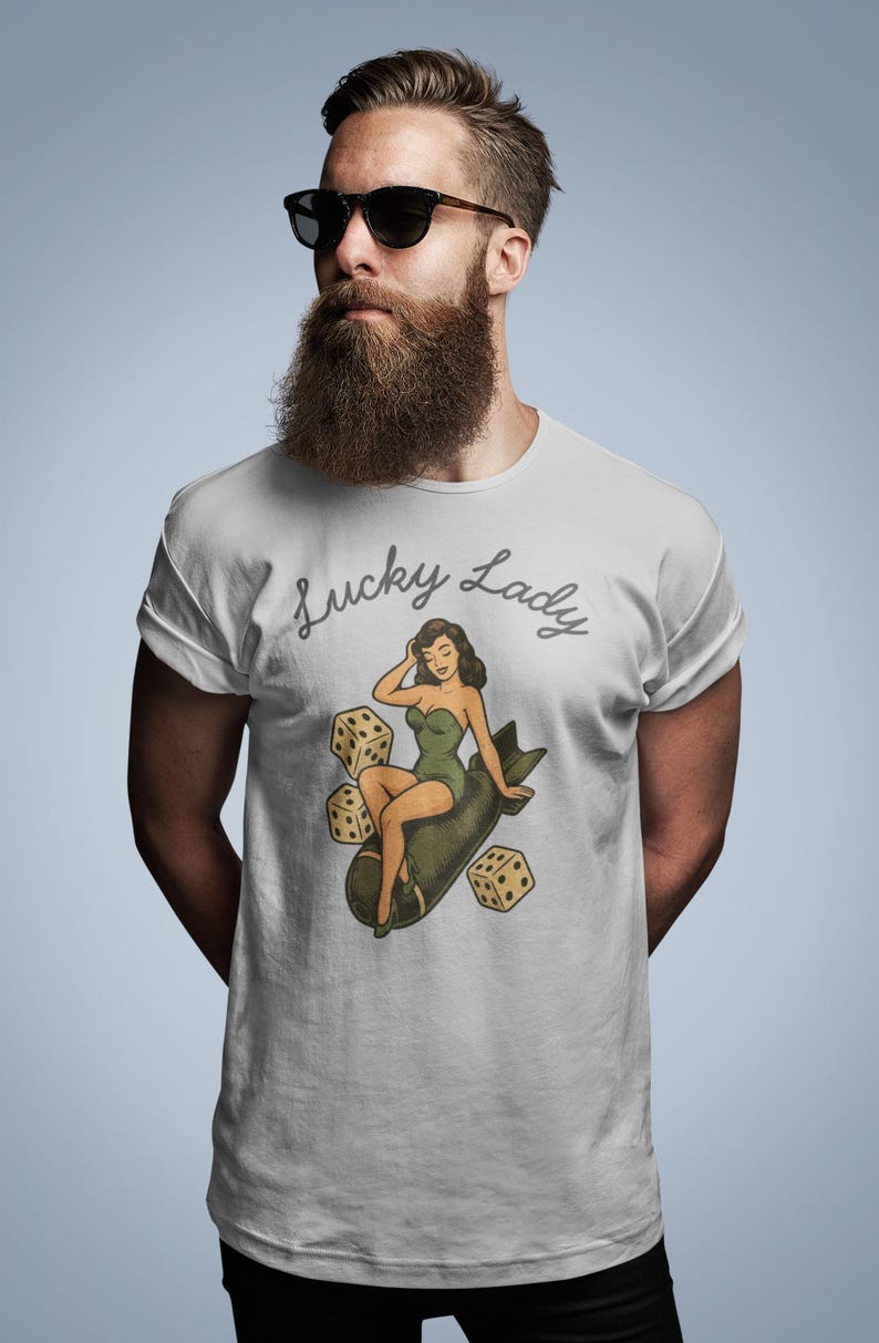 May include: A light gray short-sleeved t-shirt featuring a graphic print of a woman in a green swimsuit sitting on a dark green bomb, surrounded by three beige dice. The text Lucky Lady is printed in a dark gray cursive script above the graphic.