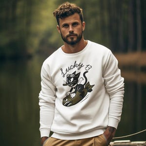 May include: White sweatshirt with a graphic of a black cat riding a bomb, with the text "Lucky 13". The cat is wearing sunglasses and smoking a cigar. The sweatshirt is paired with tan cargo pants.