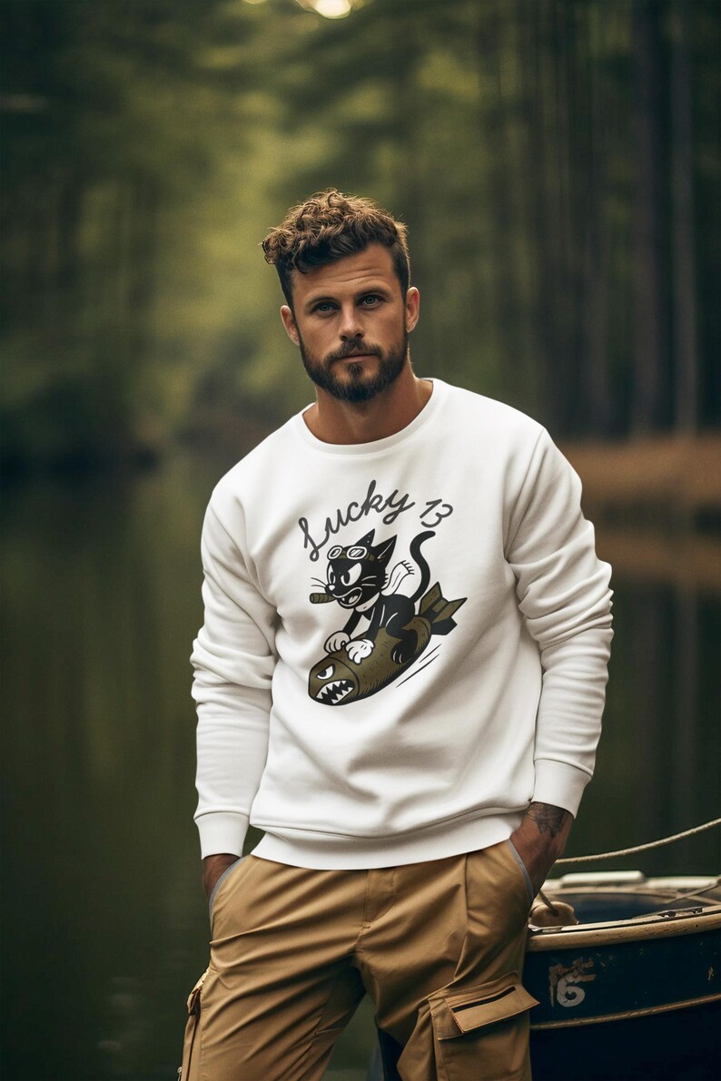 May include: White sweatshirt with a graphic of a black cat riding a bomb, with the text "Lucky 13". The cat is wearing sunglasses and smoking a cigar. The sweatshirt is paired with tan cargo pants.