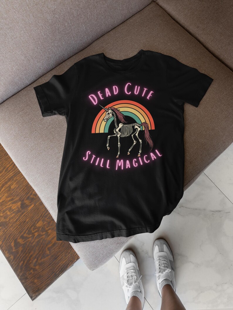May include: Black t-shirt featuring a skeletal unicorn design with the text "Dead Cute Still Magical" above and below the rainbow graphic. The unicorn has a horn and a flowing mane. The shirt is displayed on a gray surface.