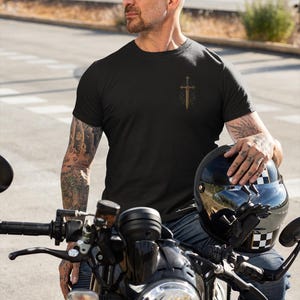 May include: Black t-shirt with a gold sword and cross design. The person is wearing blue jeans and is sitting on a black motorcycle. A black helmet with a checkered pattern is held in the person's hand.