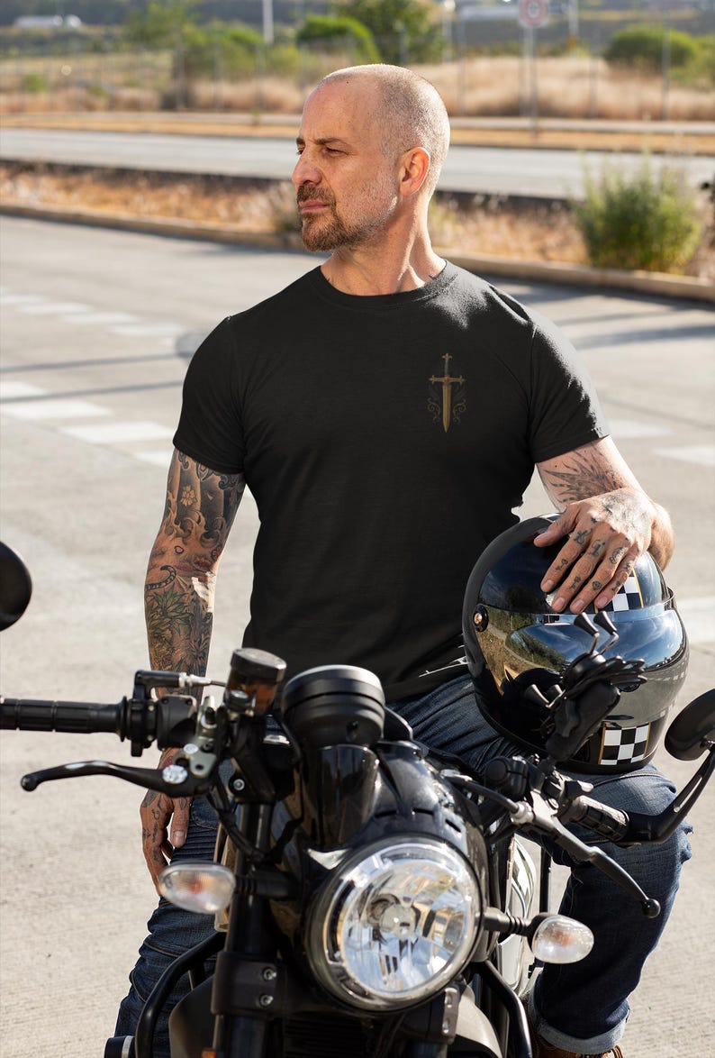 May include: Black t-shirt with a gold sword and cross design. The person is wearing blue jeans and is sitting on a black motorcycle. A black helmet with a checkered pattern is held in the person's hand.