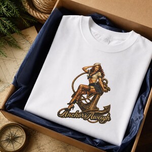 May include: A white t-shirt featuring a graphic print of a person in a sailor outfit sitting on a large metal anchor. The design includes a coiled rope and the text "Anchor's Aweigh" in a stylized font at the bottom of the graphic illustration.