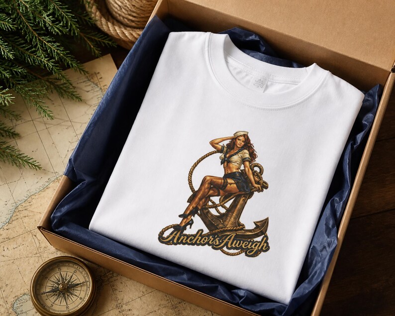May include: A white t-shirt featuring a graphic print of a person in a sailor outfit sitting on a large metal anchor. The design includes a coiled rope and the text "Anchor's Aweigh" in a stylized font at the bottom of the graphic illustration.