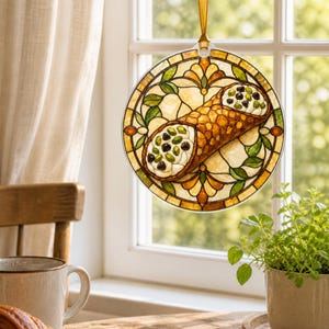 May include: A circular hanging ornament featuring a stained glass style design of a cannoli filled with cream, chocolate chips, and pistachios. The artwork is framed by amber, green, and cream geometric patterns and hangs from a gold ribbon.