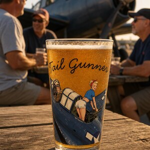May include: A clear glass pint filled with golden beer, featuring a graphic of a blue military aircraft with a pilot and a figure in a blue shirt and white shorts. The text Tail Gunner is printed in black script above the illustrated aircraft.