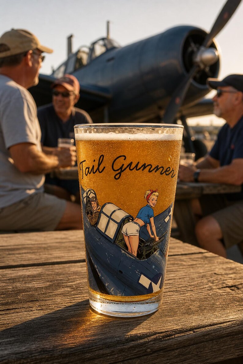 May include: A clear glass pint filled with golden beer, featuring a graphic of a blue military aircraft with a pilot and a figure in a blue shirt and white shorts. The text Tail Gunner is printed in black script above the illustrated aircraft.