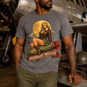 Peut inclure: Un t-shirt gris anthracite &agrave; manches courtes avec une impression graphique repr&eacute;sentant un personnage pin-up assis sur une bombe devant une pleine lune. Sous le motif, une banni&egrave;re rouge affiche le texte MIDNIGHT MISSION en lettres capitales.