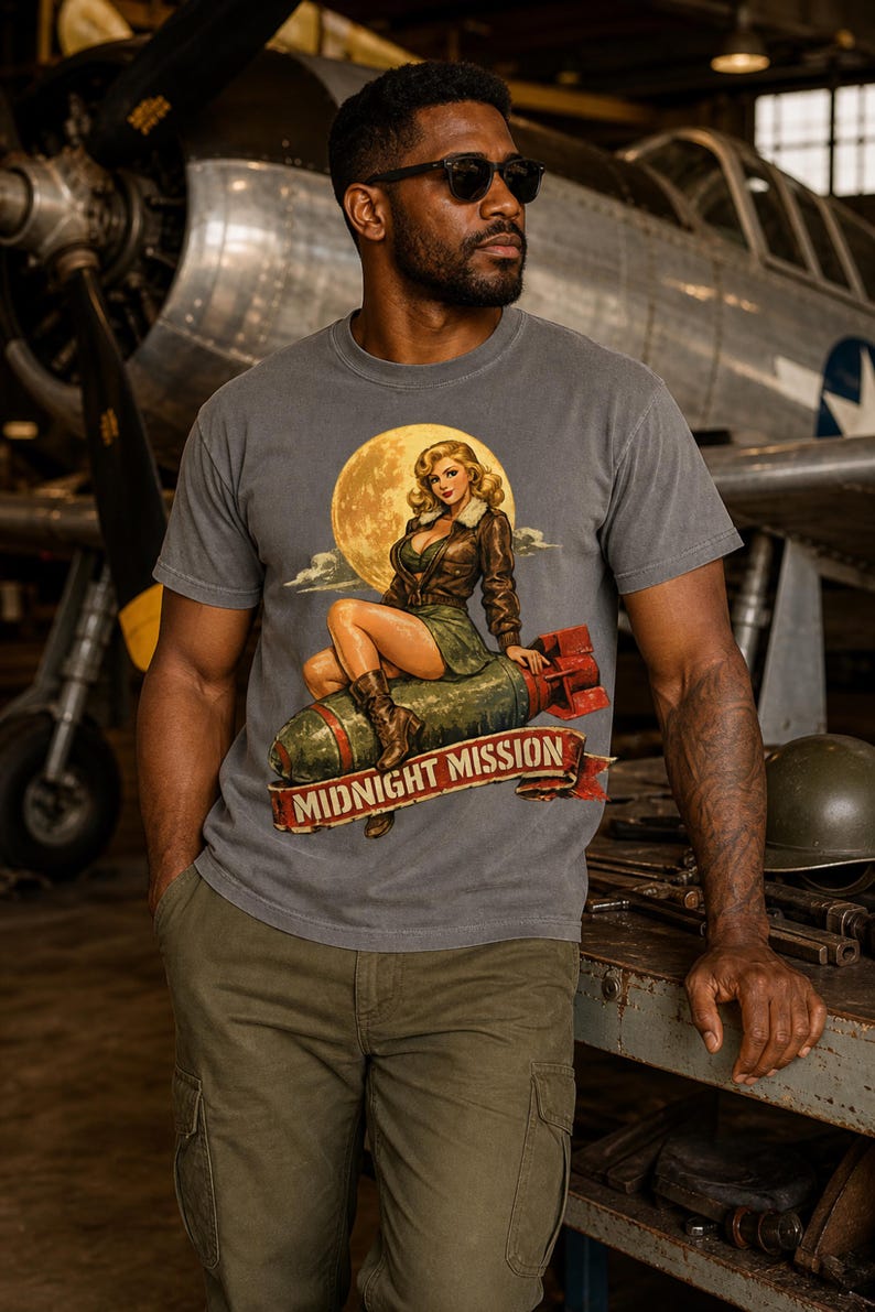 Peut inclure: Un t-shirt gris anthracite &agrave; manches courtes avec une impression graphique repr&eacute;sentant un personnage pin-up assis sur une bombe devant une pleine lune. Sous le motif, une banni&egrave;re rouge affiche le texte MIDNIGHT MISSION en lettres capitales.