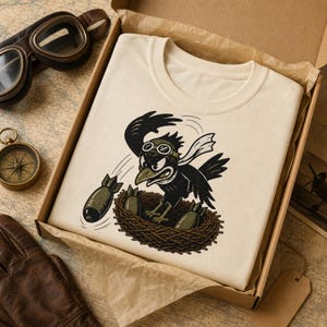 Puede incluir: Una camiseta de color crema con una ilustraci&oacute;n gr&aacute;fica de un p&aacute;jaro negro que lleva gafas de aviador y una bufanda, sentado en un nido y lanzando bombas. El p&aacute;jaro tiene una expresi&oacute;n determinada y el dise&ntilde;o es de estilo vintage.
