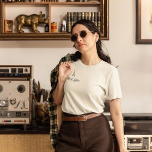 May include: A woman wearing a white t-shirt with an Eiffel Tower graphic and the text "City of Lights". She is wearing brown pants, a brown belt, and sunglasses. A plaid jacket is draped over her shoulder.