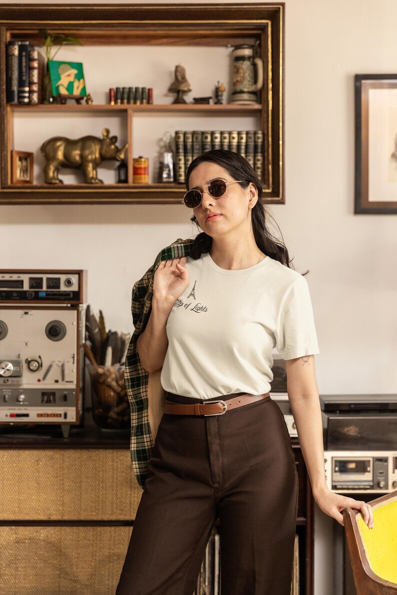 May include: A woman wearing a white t-shirt with an Eiffel Tower graphic and the text "City of Lights". She is wearing brown pants, a brown belt, and sunglasses. A plaid jacket is draped over her shoulder.