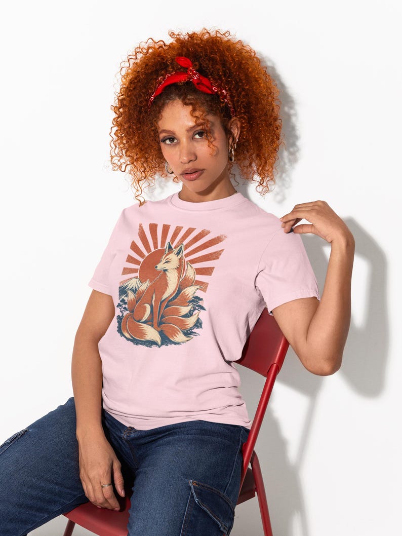 May include: A light pink short-sleeve t-shirt featuring a graphic print of a multi-tailed fox sitting in front of a stylized red sunburst. The fox is rendered in shades of orange and cream, with dark blue accents at the base of the design.