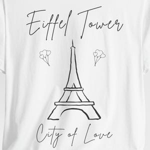 May include: White t-shirt featuring a black line drawing of the Eiffel Tower with the words "Eiffel Tower" above and "City of Love" below. Two small heart-shaped balloon clusters are on either side of the tower.