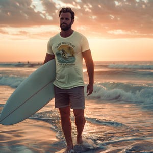 May include: A person walking out of the ocean, holding a surfboard. They are wearing a light-colored t-shirt with a graphic that says "Saint Augustine Catch the coastline Est. 1988". The sky is a warm orange and the ocean is a deep blue.