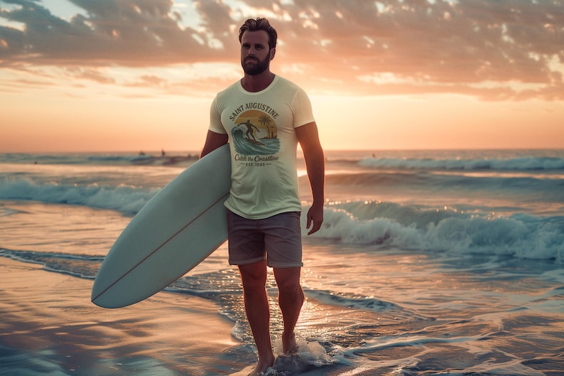 May include: A person walking out of the ocean, holding a surfboard. They are wearing a light-colored t-shirt with a graphic that says "Saint Augustine Catch the coastline Est. 1988". The sky is a warm orange and the ocean is a deep blue.