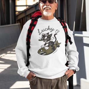 May include: White sweatshirt with a graphic of a black cat wearing goggles and smoking a cigar, riding a bomb. The text "Lucky 13" is above the cat. The person is wearing a white cap, sunglasses, and cargo pants.