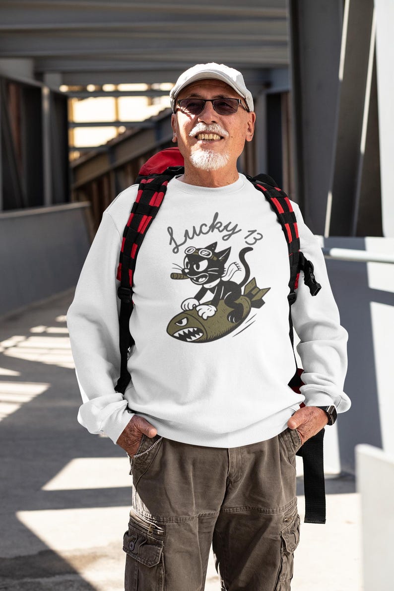 May include: White sweatshirt with a graphic of a black cat wearing goggles and smoking a cigar, riding a bomb. The text "Lucky 13" is above the cat. The person is wearing a white cap, sunglasses, and cargo pants.