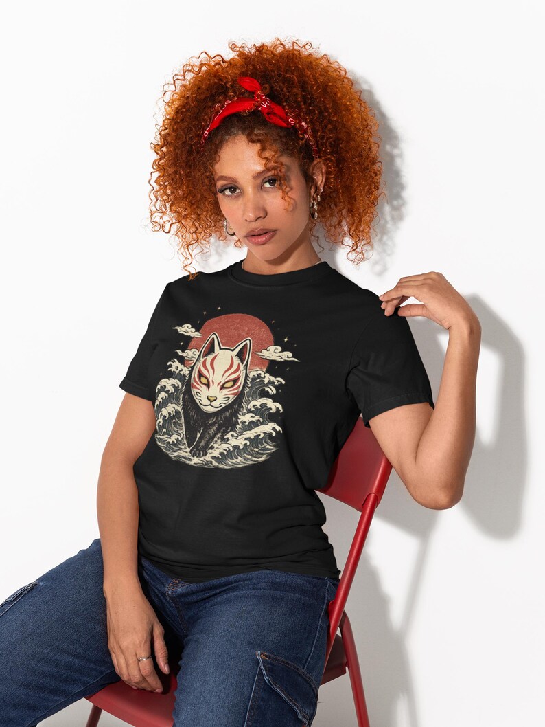 May include: A black t-shirt featuring a graphic print of a fox mask with red and white markings, positioned above stylised ocean waves and a large red sun. The design includes small white cloud accents surrounding the central fox mask illustration.