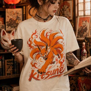 K&ouml;nnte beinhalten: Ein cremefarbenes Kurzarm-T-Shirt mit einem gro&szlig;en Grafikdruck eines orangefarbenen neunschw&auml;nzigen Fuchses, der von stilisierten Flammen umgeben ist. Das Wort Kitsune ist in einer kr&auml;ftigen Pinselstrich-Schrift in Rot und Orange abgebildet.