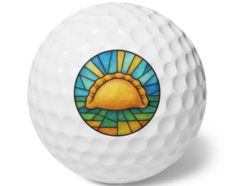 May include: A white golf ball featuring a circular graphic in the center. The graphic depicts a golden-brown empanada centered against a stained-glass style background with radiating blue, yellow, and green geometric segments enclosed in a black border.