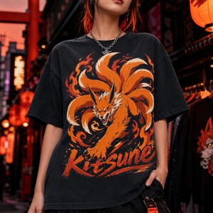 K&ouml;nnte beinhalten: Ein schwarzes Kurzarm-T-Shirt mit einem Grafikdruck eines orangefarbenen neunschw&auml;nzigen Fuchses, der von stilisierten Flammen umgeben ist. Das Wort Kitsune ist unter der Fuchsillustration in einer fetten Pinselstrich-Schrift gedruckt.