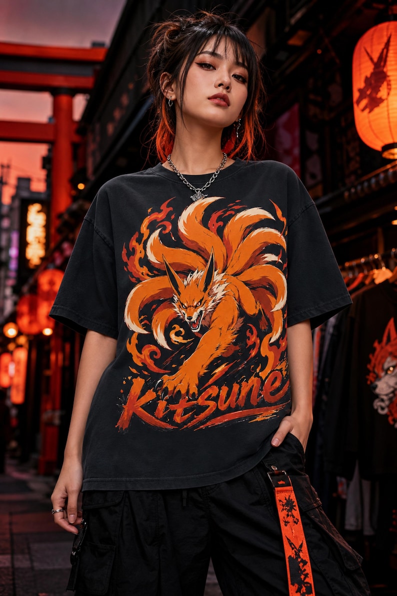 K&ouml;nnte beinhalten: Ein schwarzes Kurzarm-T-Shirt mit einem Grafikdruck eines orangefarbenen neunschw&auml;nzigen Fuchses, der von stilisierten Flammen umgeben ist. Das Wort Kitsune ist unter der Fuchsillustration in einer fetten Pinselstrich-Schrift gedruckt.