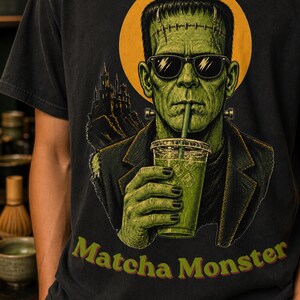 Może przedstawiać: Czarny t-shirt z grafiką przedstawiającą zielonego potwora w okularach przeciwsłonecznych, pijącego zielony nap&oacute;j z plastikowego kubka przez słomkę. Za postacią znajduje się ż&oacute;łte koło i zamek. Poniżej znajduje się napis Matcha Monster.